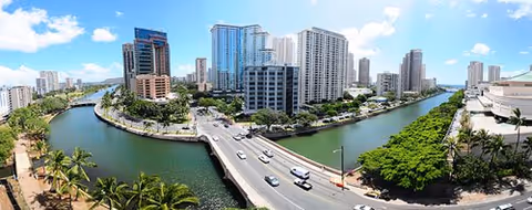 Panoramic view of a cityscape featuring a river curving through the area, surrounded by tall buildings, a bridge with vehicles crossing, and lush greenery along the riverbanks under a partly cloudy sky.