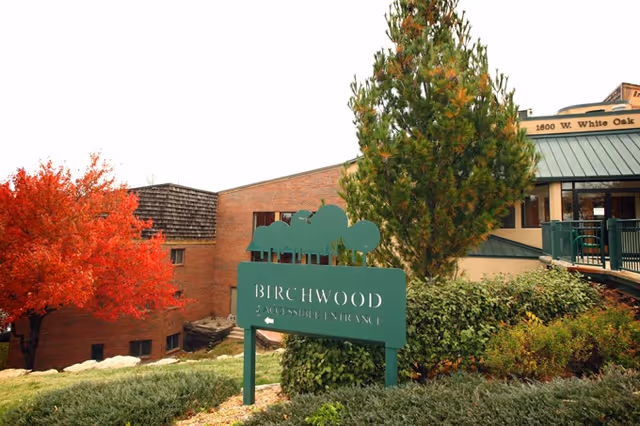 Green Birchwood sign amid landscaping in front of a senior living building with autumn trees and the entrance visible.