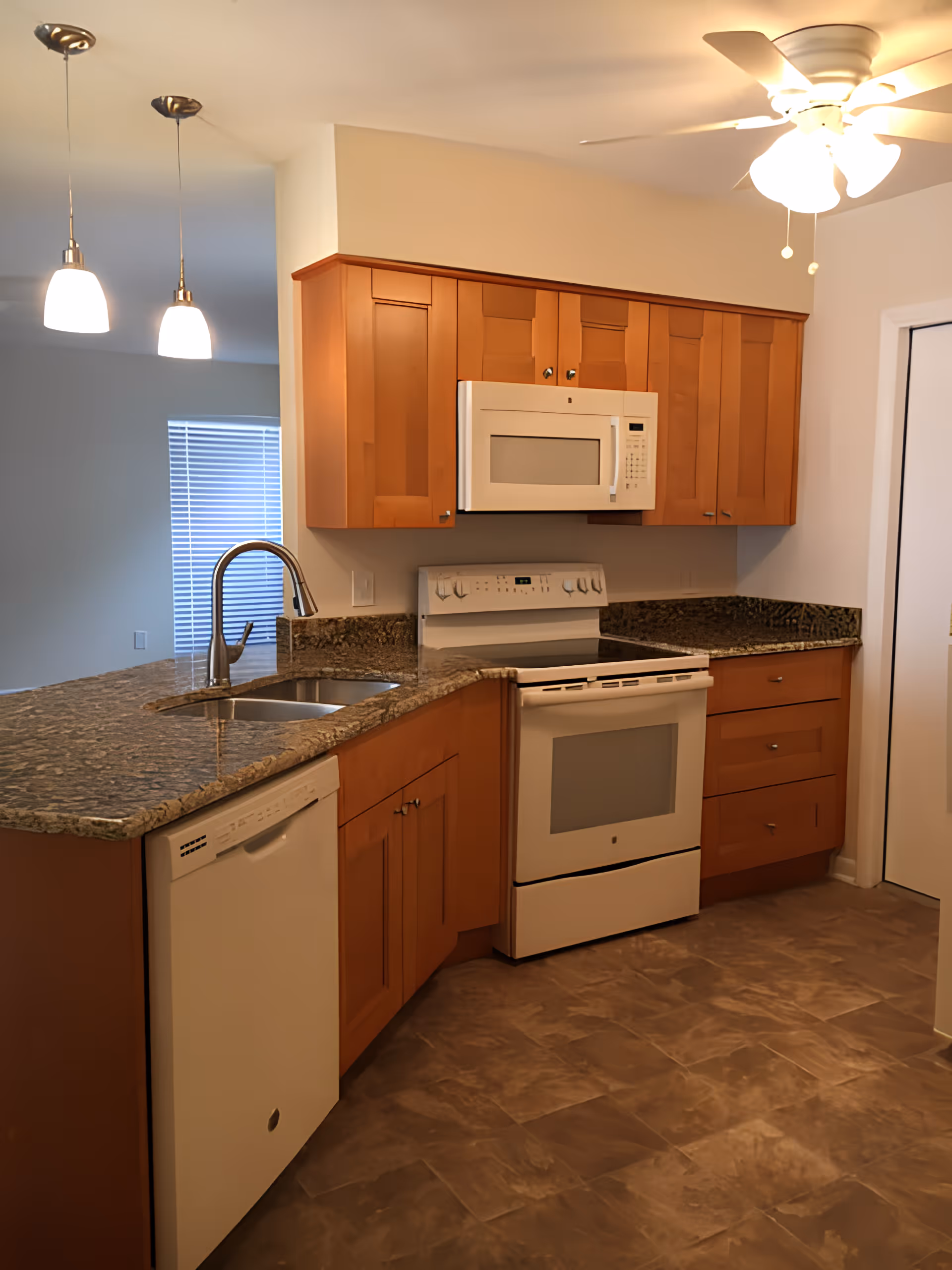 Modern kitchen with wooden cabinets, granite countertops, a white dishwasher, a white electric stove with oven, a white microwave above the stove, a double sink with a curved faucet, two hanging pendant lights, and a ceiling fan with lights.
