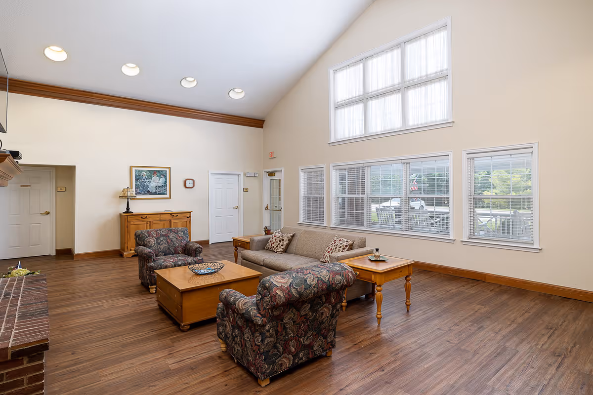 A spacious living room with high vaulted ceilings and large windows letting in natural light. The room features a beige sofa with patterned pillows, two floral upholstered armchairs, a wooden coffee table, and side tables. There is a wooden cabinet against the wall with a lamp and a framed picture above it. The floor is wood, and there is a brick fireplace partially visible on the left side.