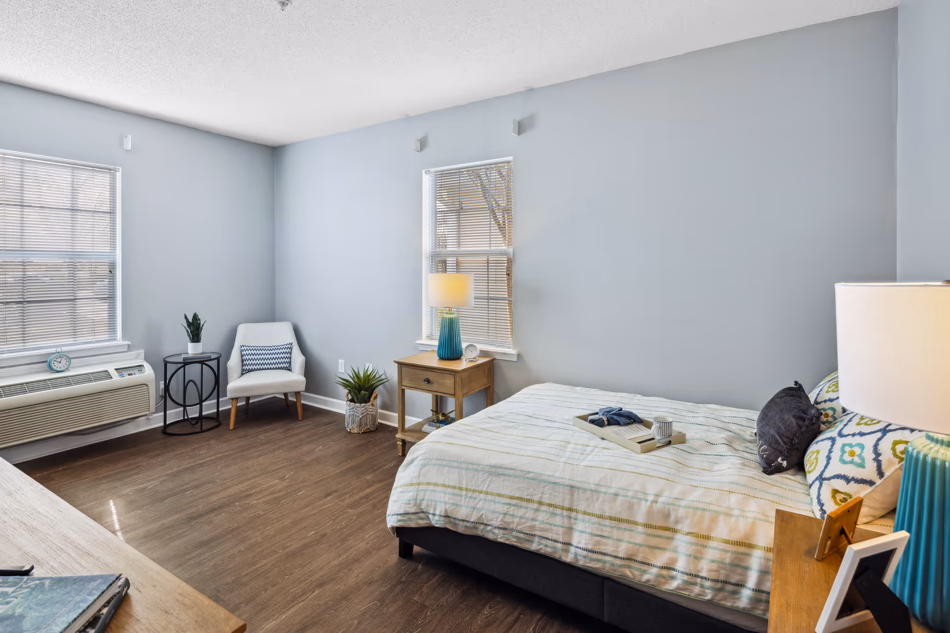 A bright and clean bedroom with light blue walls, two windows with blinds, a bed with patterned pillows and a striped bedspread, a wooden nightstand with a blue lamp, a small round table with a plant, a white cushioned chair with a decorative pillow, and wooden flooring.