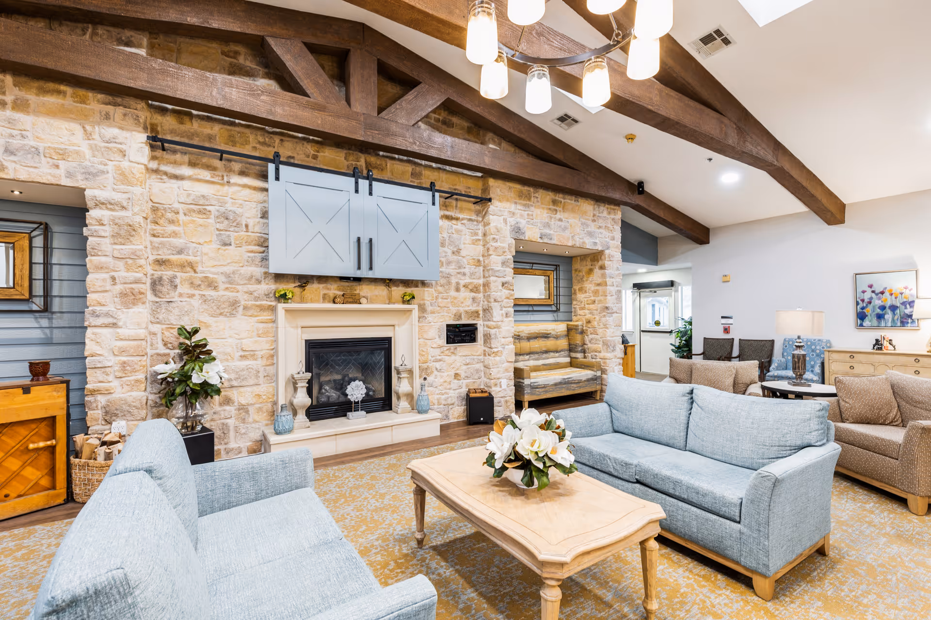 Cozy communal living room with light-blue sofas facing a stone fireplace beneath exposed wooden beams and a central coffee table with flowers.