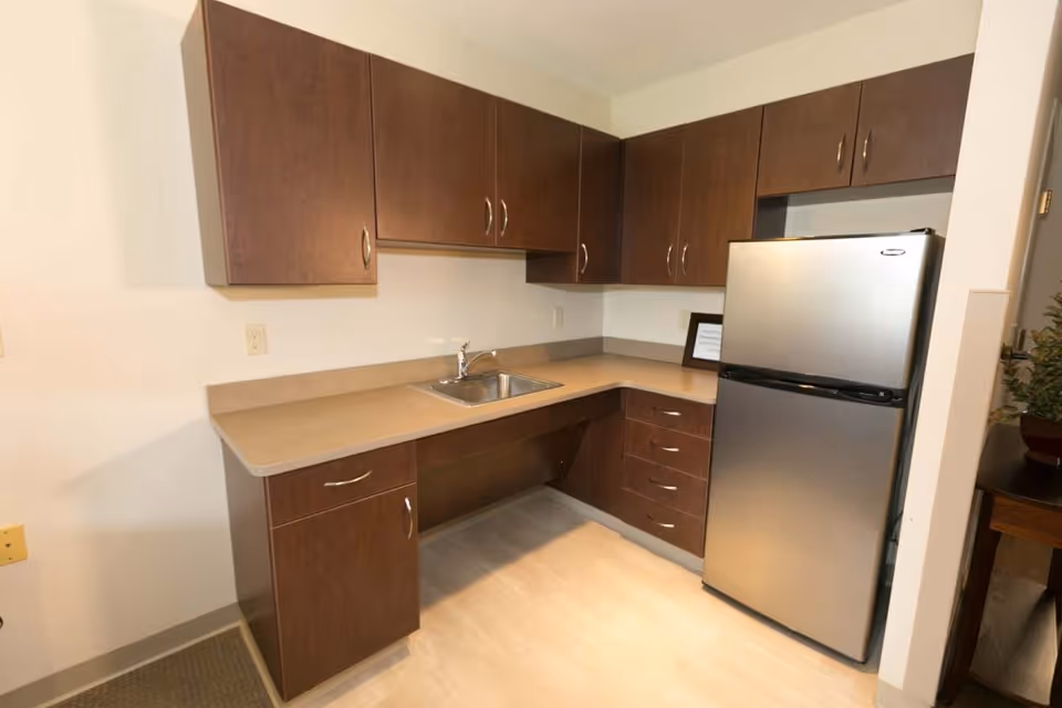 A small kitchen area with brown wooden cabinets, a beige countertop, a stainless steel sink, and a stainless steel refrigerator. The floor is light-colored, and there is a small framed item on the countertop near the refrigerator.