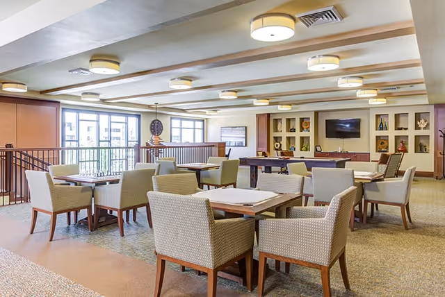 A spacious, well-lit common area with multiple tables and cushioned chairs arranged for socializing or activities. The room features large windows letting in natural light, ceiling lights, a TV mounted on the wall, and built-in shelves with decorative items.
