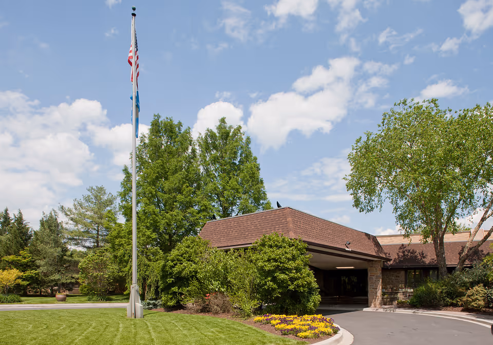 Exterior view of Stonegates facility entrance with a covered driveway, surrounded by green trees, bushes, and a flower bed. A flagpole with an American flag and another flag is visible on the left side under a partly cloudy sky.