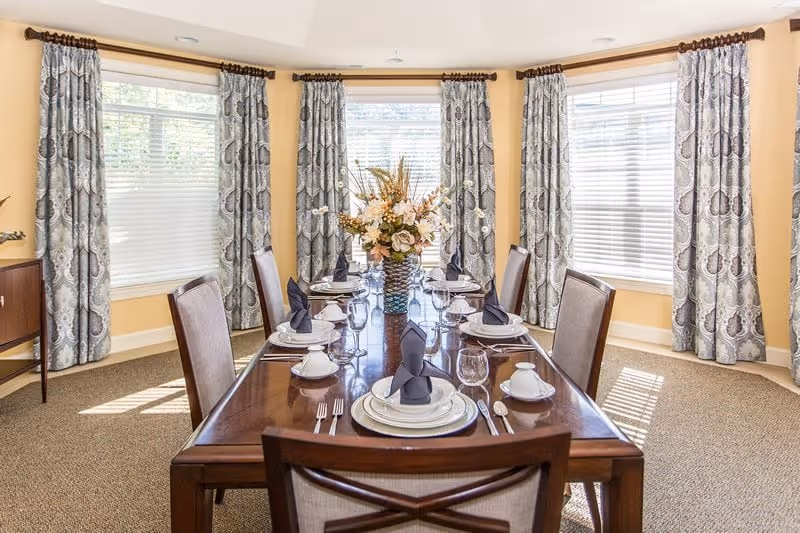 Sunlit dining room with a long wooden table set for eight with plates, folded napkins, glassware, upholstered chairs, patterned curtains, and a floral centerpiece.