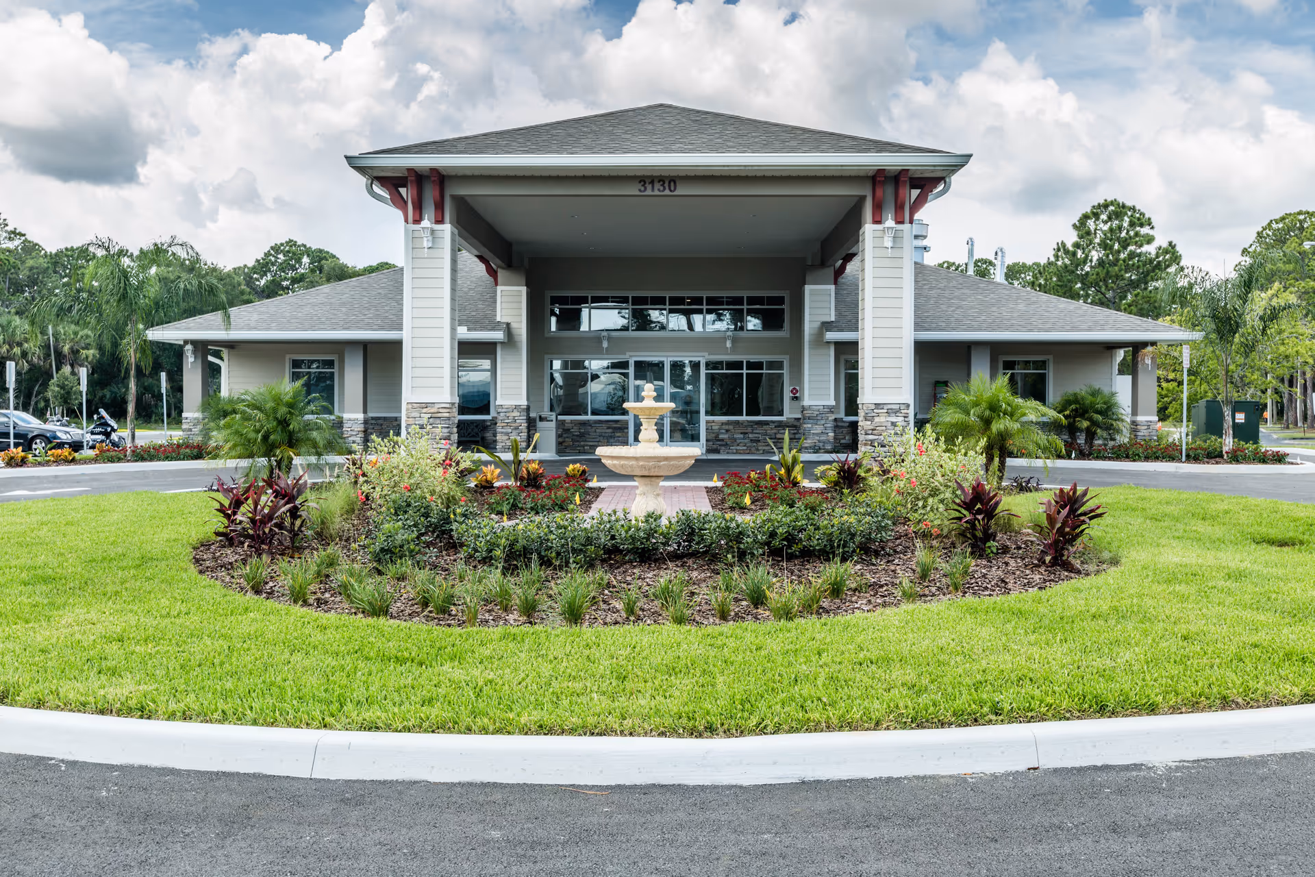 Front exterior view of a single-story building with a covered entrance, stone accents on the lower walls, and a landscaped circular garden with a fountain in the center. The building has the number 3130 above the entrance and is surrounded by green grass and trees under a partly cloudy sky.