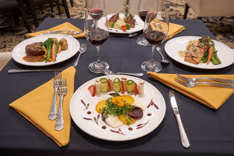 A dining table set with four plates of gourmet food, each accompanied by a glass of red wine. The table has a black tablecloth and yellow napkins with silver cutlery arranged neatly. The dishes include a steak with vegetables, a salmon fillet with asparagus and carrots, a plate with rolled cucumber and strawberries, and a lamb dish garnished with rosemary and cherry tomatoes.