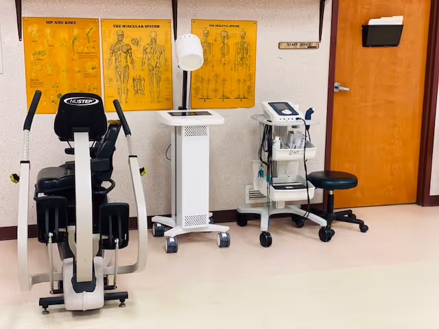 A medical or physical therapy room with exercise equipment including a NuStep machine, medical devices on wheeled carts, anatomical posters of the muscular system and hip and knee on the wall, a wooden door labeled 'Staff Office', and a black stool.