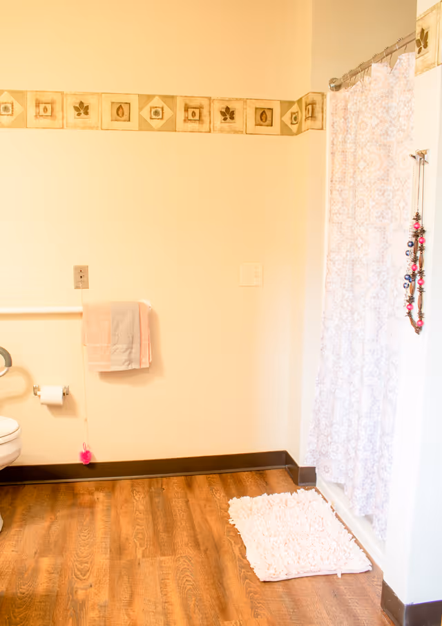 A bathroom with a wooden floor, a white toilet partially visible on the left, a towel rack with a pink and gray towel, a toilet paper holder, a white and pink bath mat on the floor, and a shower with a patterned curtain and hanging beaded decoration on the right. The walls are light-colored with a decorative border near the ceiling.