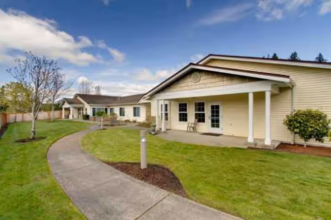 Curved concrete walkway leads to a single-story beige senior living building with green lawn and porch.
