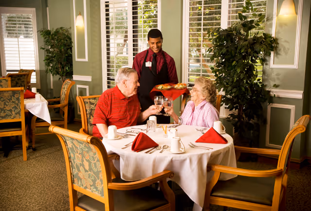 An elderly man and woman sitting at a round dining table in a well-lit dining room, clinking their glasses in a toast while a waiter in a red shirt and black apron serves food. The table is set with white tablecloth, red napkins, cups, and silverware. The room has green walls, large windows with white shutters, and decorative plants.