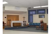 Interior view of a chapel or small worship area with an altar covered by a white cloth, a wooden cross on the wall, a lectern, blue chairs arranged in rows, and a floral arrangement near a wooden table.