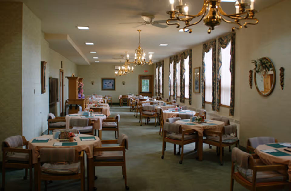 Interior view of a retirement home dining room with multiple round tables covered with beige tablecloths and green placemats. Each table is surrounded by wooden chairs with cushions. The room has several windows with patterned curtains, chandeliers hanging from the ceiling, and framed artwork on the walls.