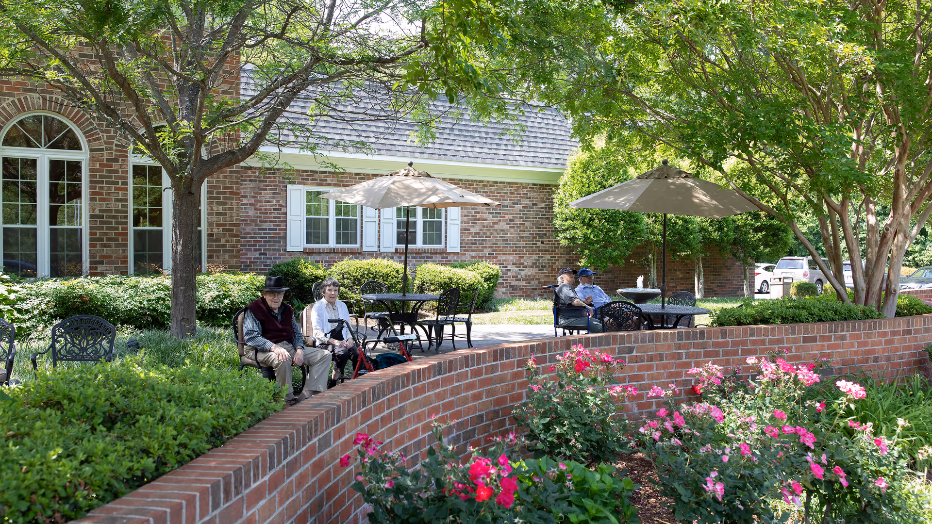 Outdoor patio area at Paul Spring Retirement Community with elderly residents sitting on benches and chairs under large umbrellas. The space is surrounded by greenery, trees, and blooming pink flowers, with a brick building in the background.
