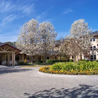Outdoor view of The Village at Rancho Solano facility showing a circular driveway with landscaped flower beds and trees with white blossoms under a clear blue sky. The building is visible in the background with multiple windows and a covered entrance area.