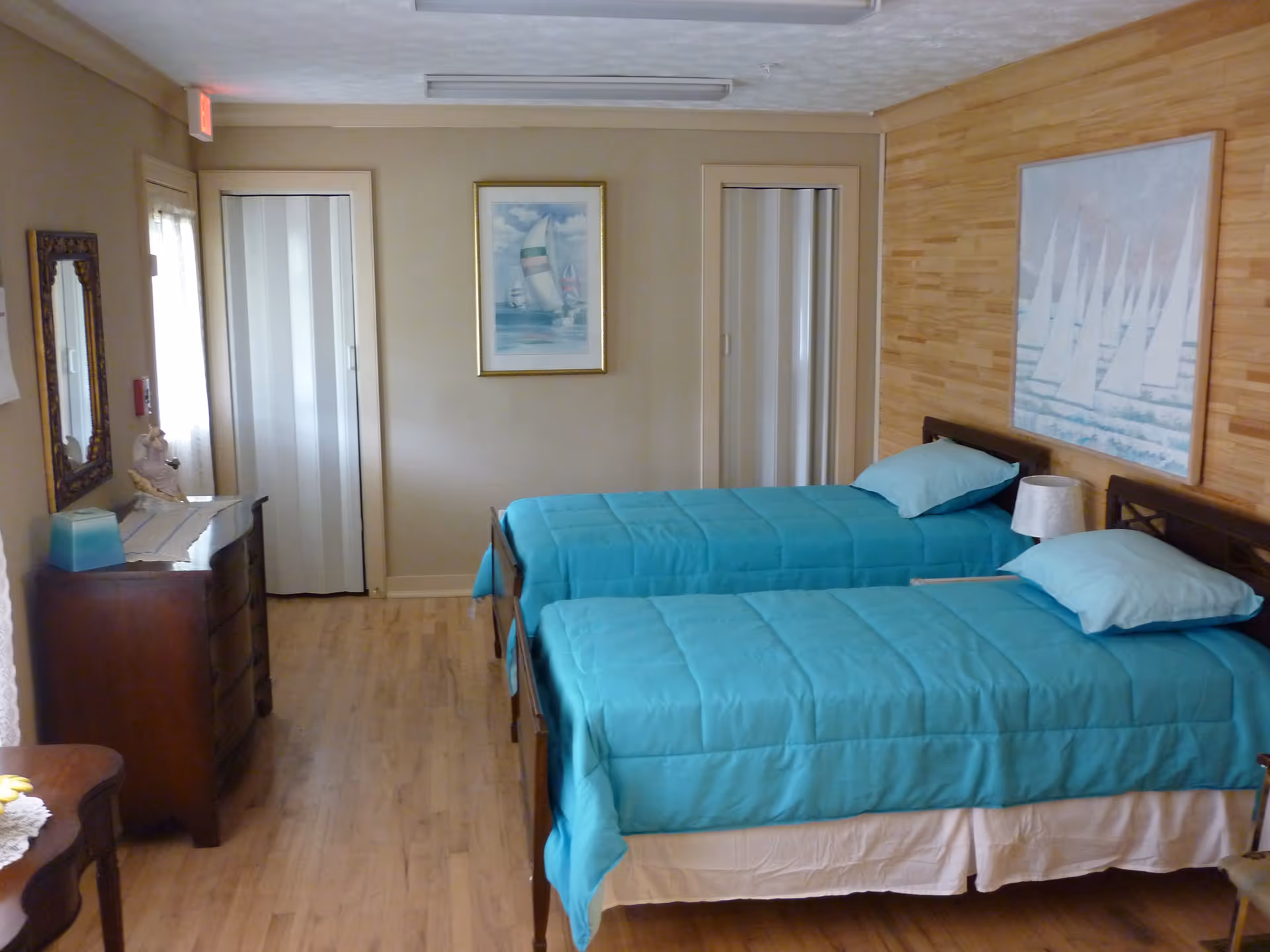 A bedroom with two twin beds covered in turquoise bedding and matching pillows. The room has wooden flooring and light-colored walls, with a large painting of sailboats above the beds. There is a wooden dresser with a mirror on the left side and two closed doors on the far wall.