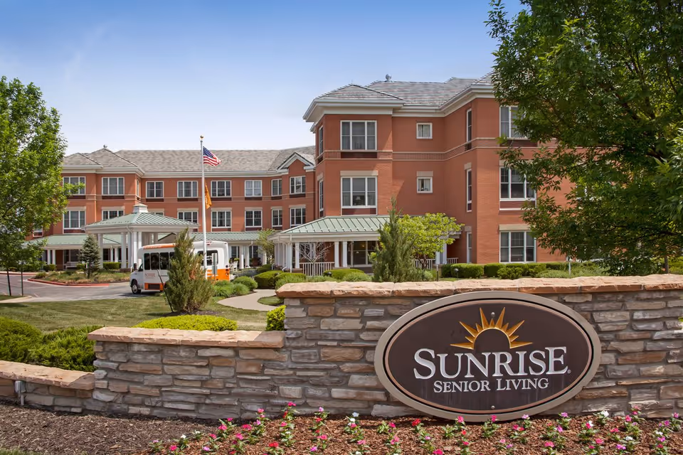 Exterior view of Sunrise Senior Living facility showing a multi-story brick building with many windows, a covered entrance with a driveway, a shuttle bus parked nearby, landscaped greenery, and a stone sign with the Sunrise Senior Living logo in front.
