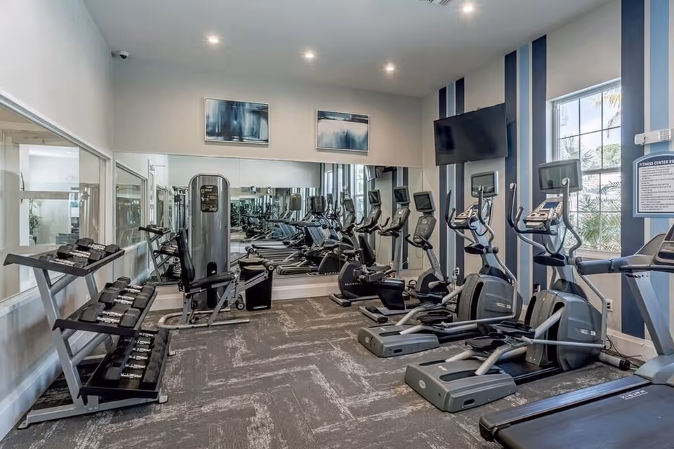 Interior view of a fitness center with exercise equipment including elliptical machines, treadmills, weight machines, and a rack of dumbbells. The room has large mirrors on one wall, striped blue and white walls, and windows letting in natural light.