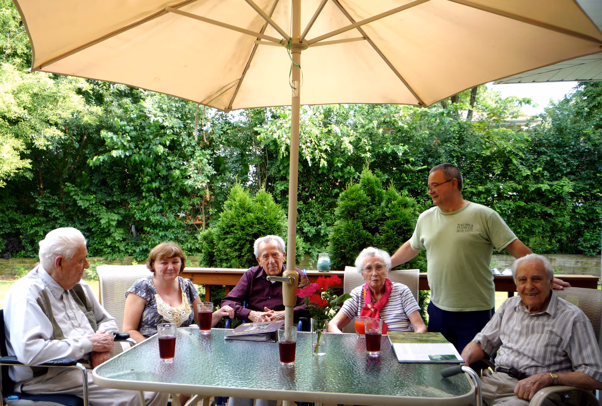 A group of five elderly people and one middle-aged man sitting and standing around a glass outdoor table under a large beige umbrella. The table has several glasses with red drinks and a small vase with flowers. The background shows lush green trees and bushes.