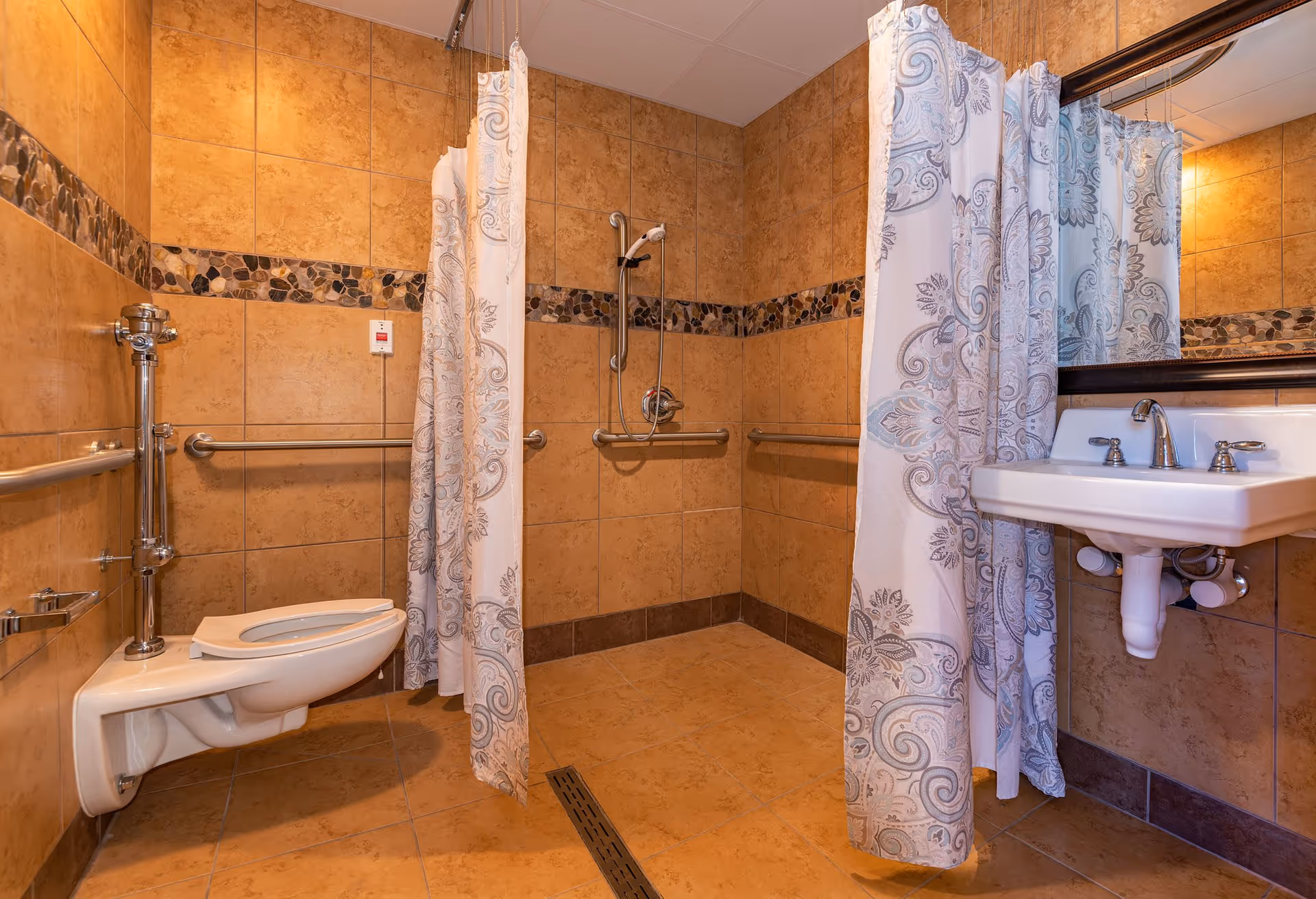 Accessible tiled bathroom with a wall-mounted toilet, grab bars, roll-in shower with curtain, and a sink with mirror.