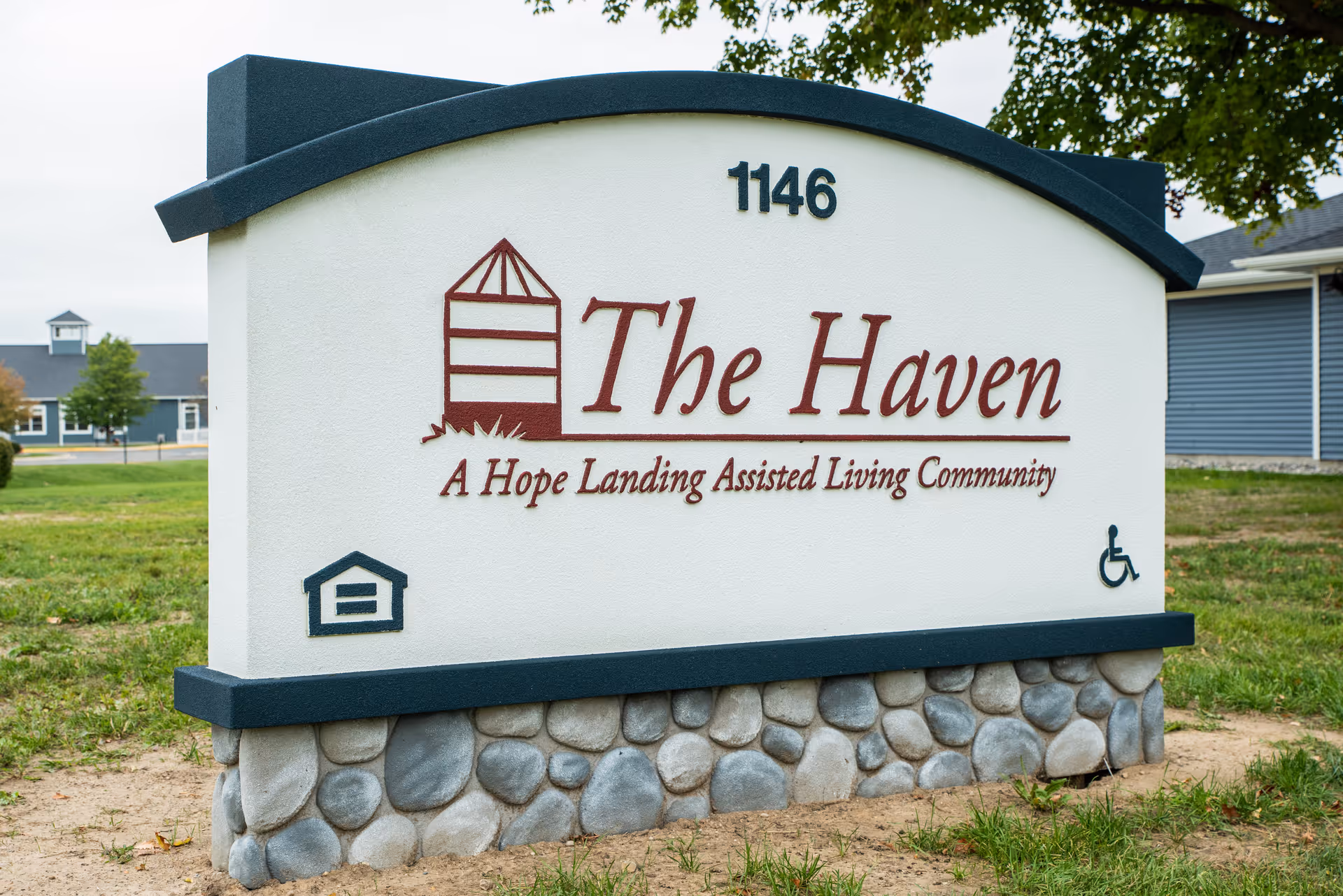 Outdoor sign for The Haven, a Hope Landing Assisted Living Community, with the address number 1146. The sign features a small house icon and a wheelchair accessibility symbol, set on a stone base with grass and buildings in the background.