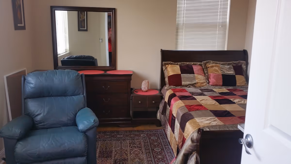 A cozy bedroom with a dark wooden bed frame and a colorful patchwork quilt. There are matching pillows on the bed. Next to the bed is a wooden nightstand with a pink Himalayan salt lamp. A wooden dresser with a large mirror above it is positioned against the wall. A blue leather recliner chair is placed near the dresser. The room has beige walls, a window with blinds, and a patterned area rug on the floor.