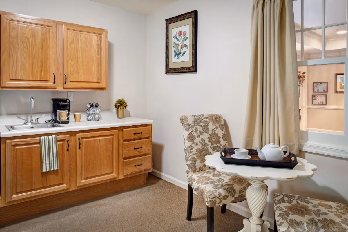 Small kitchenette and seating area with oak cabinets, a sink, patterned chairs and a small table set with a teapot.