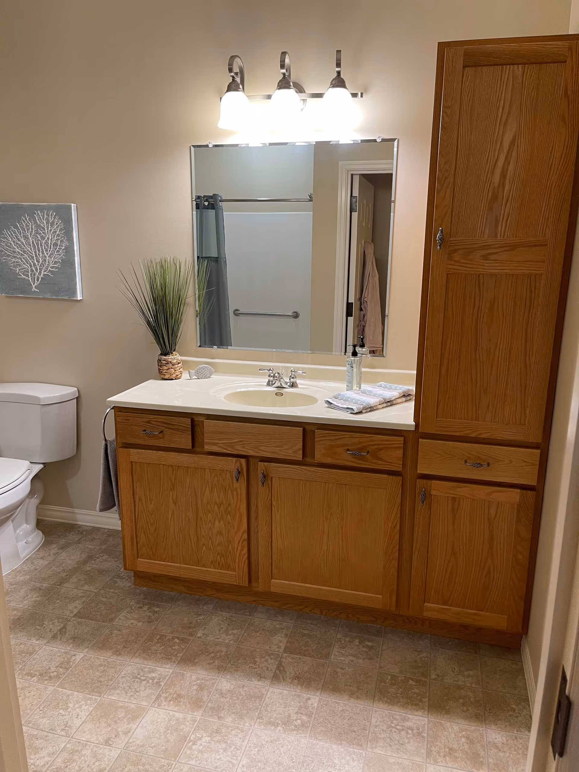 A bathroom with a wooden vanity featuring a white countertop and sink, a large mirror above it with three light fixtures, a toilet to the left, a tall wooden cabinet to the right, a small plant on the countertop, and a towel hanging on a ring beside the toilet. The floor has beige tiles and a wall decoration with a coral design is visible above the toilet.
