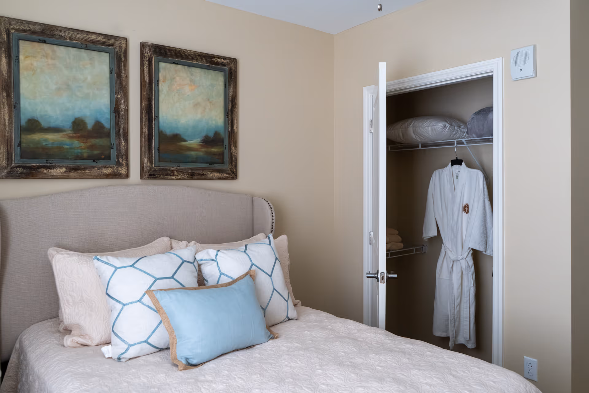 A bedroom with a beige upholstered headboard, a bed with multiple pillows including two with a blue geometric pattern and one solid blue pillow. Above the bed are two framed landscape paintings. To the right, an open closet door reveals a white bathrobe hanging inside along with folded towels and pillows on the shelf.