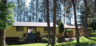 Single-story yellow building with a dark roof, nestled among tall trees and a green lawn.