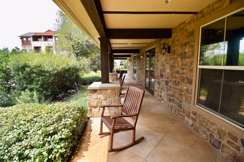 Covered outdoor patio area with stone walls and tiled floor, featuring wooden rocking chairs and surrounded by green bushes and trees.