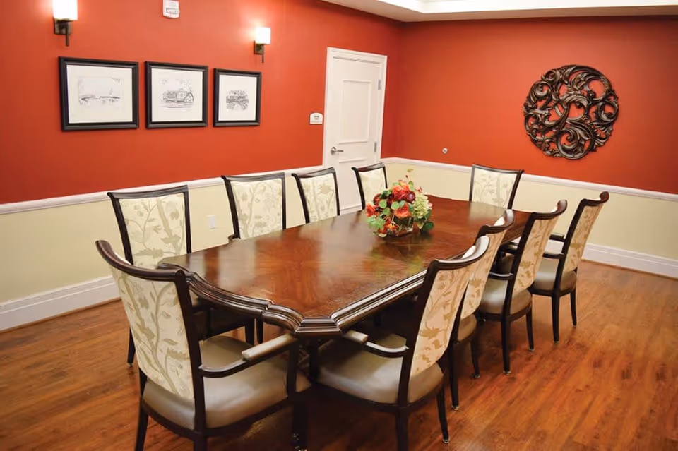 A formal dining room with a long wooden table surrounded by ten upholstered chairs with floral patterned backs. The walls are painted red on the upper half and cream on the lower half, decorated with three framed sketches and a decorative metal wall art piece. The floor is wooden, and two wall sconces provide lighting.