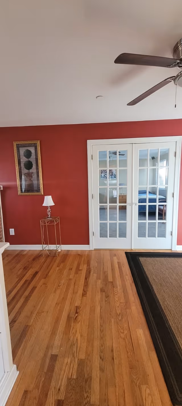 Interior view of a room with wooden flooring, a red painted wall with a framed artwork, a small table with a lamp, and white French doors leading to a bedroom with a bed and dresser visible through the glass panels. A ceiling fan is partially visible at the top right.