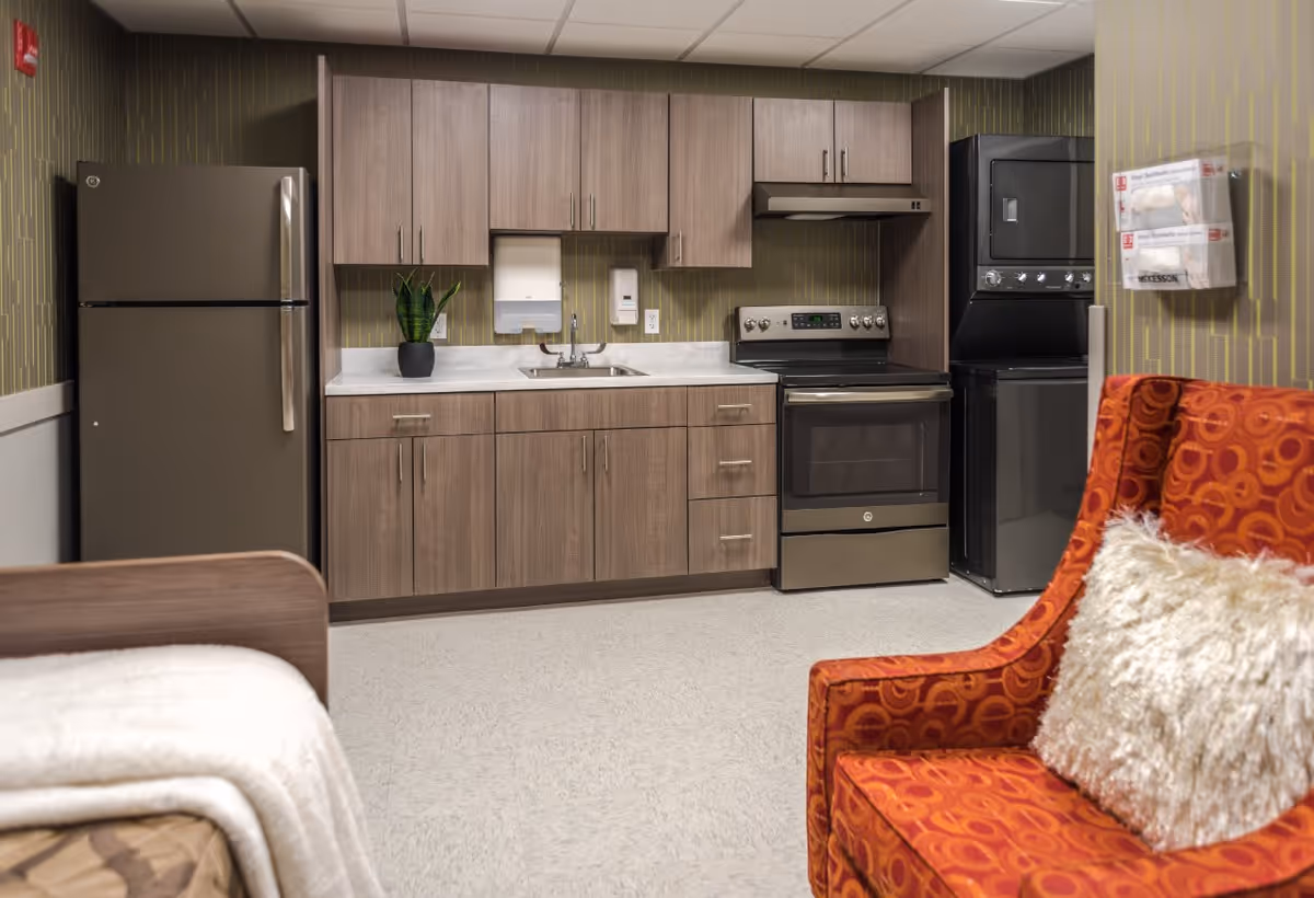 Interior view of a room featuring a kitchenette with wooden cabinets, a stainless steel refrigerator, stove, and a stacked washer and dryer. There is a small potted plant on the countertop, a sink with a soap dispenser and paper towel holder above it. In the foreground, there is an orange patterned armchair with a fluffy white pillow and part of a bed with a beige blanket is visible.