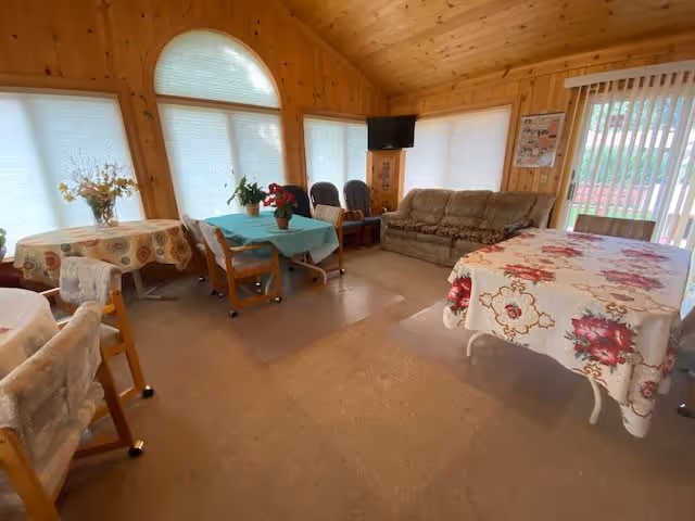 A cozy common area with wooden walls and ceiling, featuring several tables covered with floral and patterned tablecloths, chairs, a sofa, a small TV mounted on a wooden stand, and large windows with blinds letting in natural light.