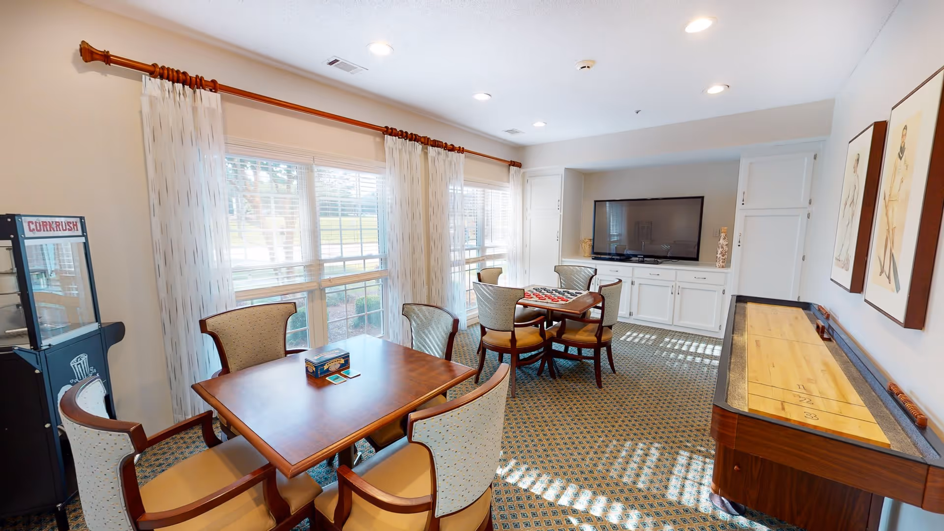 A bright and cozy common room with large windows covered by sheer curtains, a wooden table with four upholstered chairs, a shuffleboard table, a television mounted on a white cabinet, and a popcorn machine in the corner.
