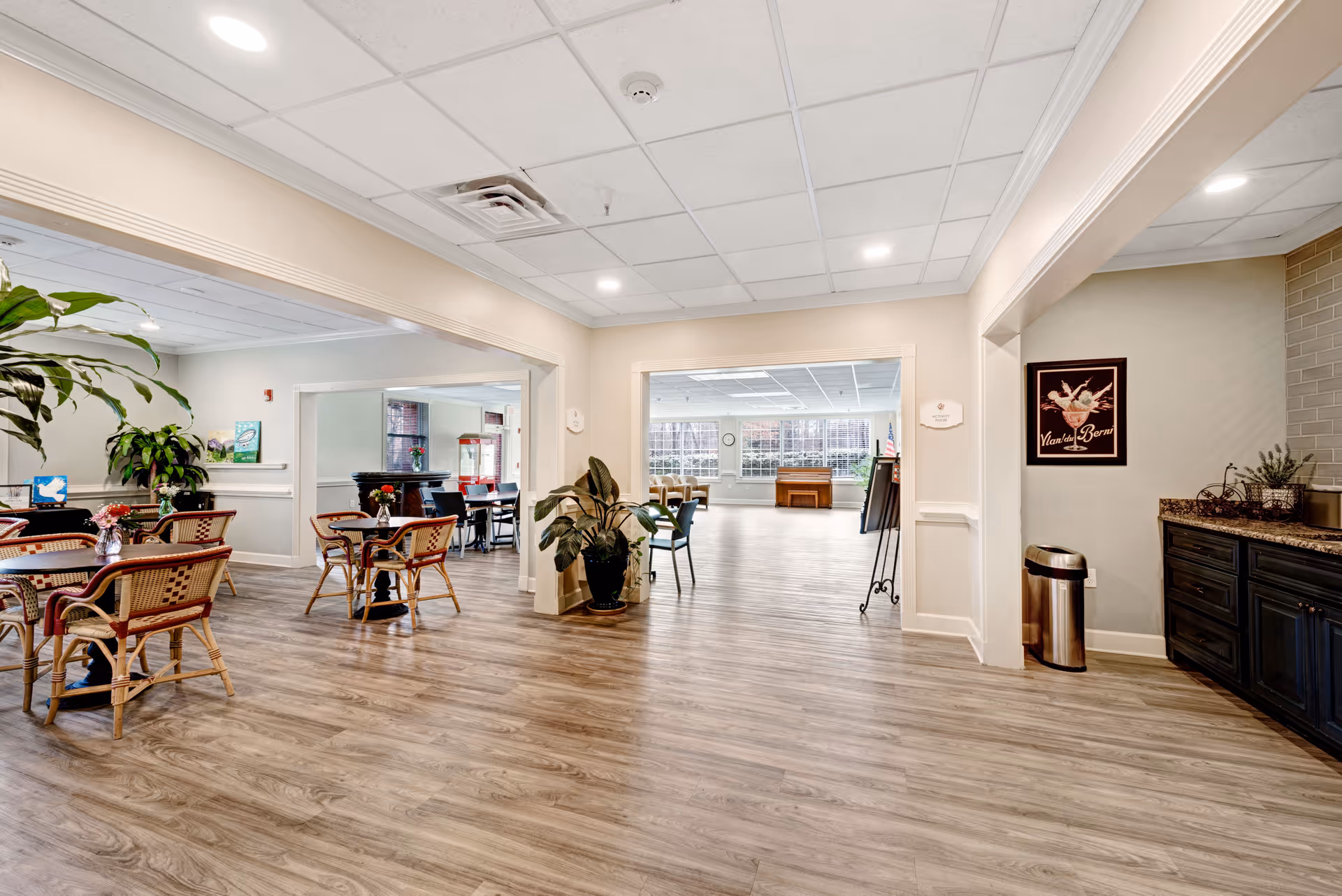 A spacious, well-lit common area in a senior living facility with wood-style flooring, several tables and chairs arranged for dining or socializing, large windows letting in natural light, potted plants, and a small counter area with a trash can and decorative items.