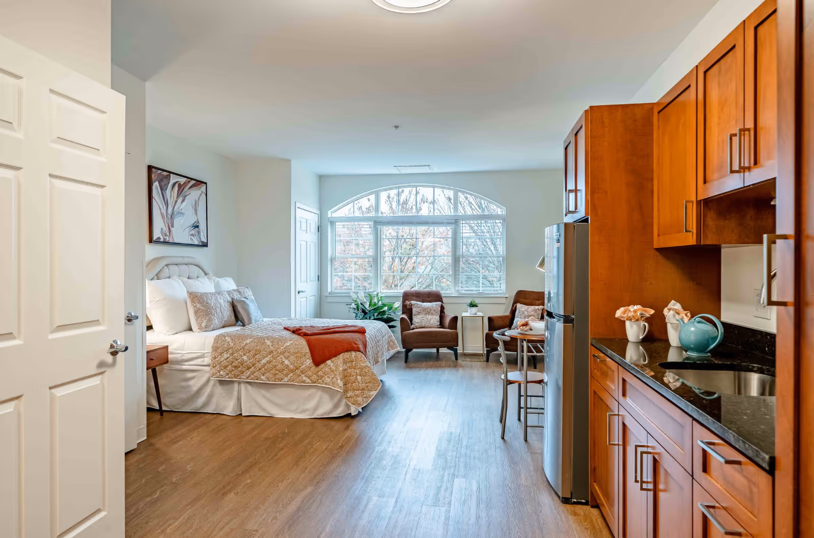 A bright studio apartment with a bed on the left side, two brown armchairs with a small table near a large arched window, and a kitchenette with wooden cabinets and a black countertop on the right side.