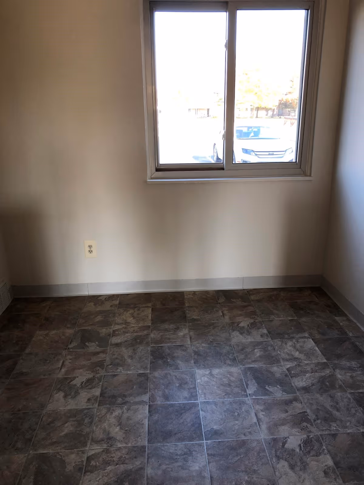 Empty room with tiled floor, beige walls, a window showing a parked car outside, and an electrical outlet on the wall.