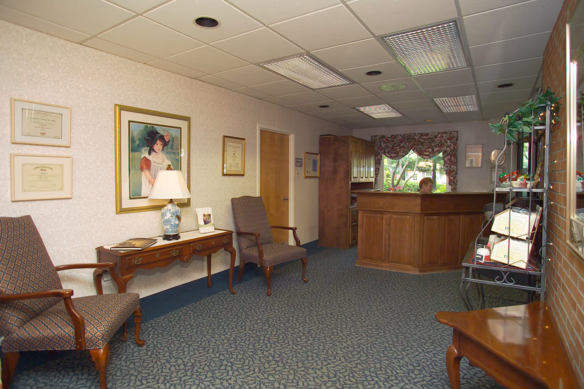 Reception area of a nursing home with upholstered chairs, a wooden front desk, framed artwork, and a lamp on a console table.