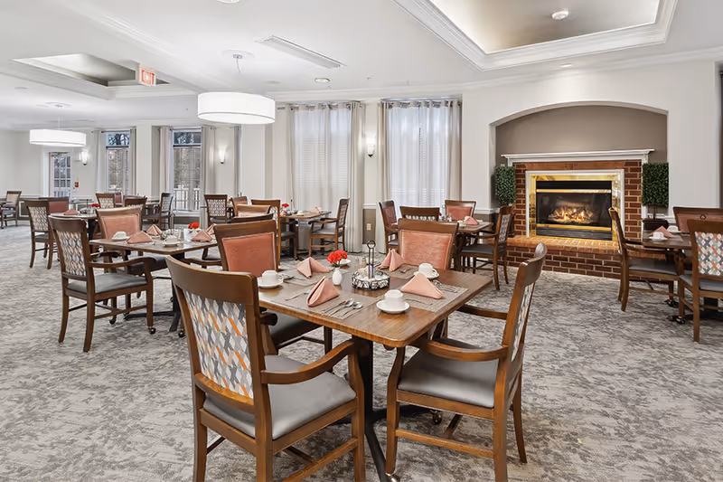 A spacious dining room with multiple wooden tables and chairs arranged neatly. Each table is set with cups, plates, silverware, and folded napkins. There is a lit fireplace with a brick surround on the right side of the room, flanked by two small green plants. Large windows with sheer curtains allow natural light to fill the room. The ceiling features recessed lighting and modern hanging light fixtures.