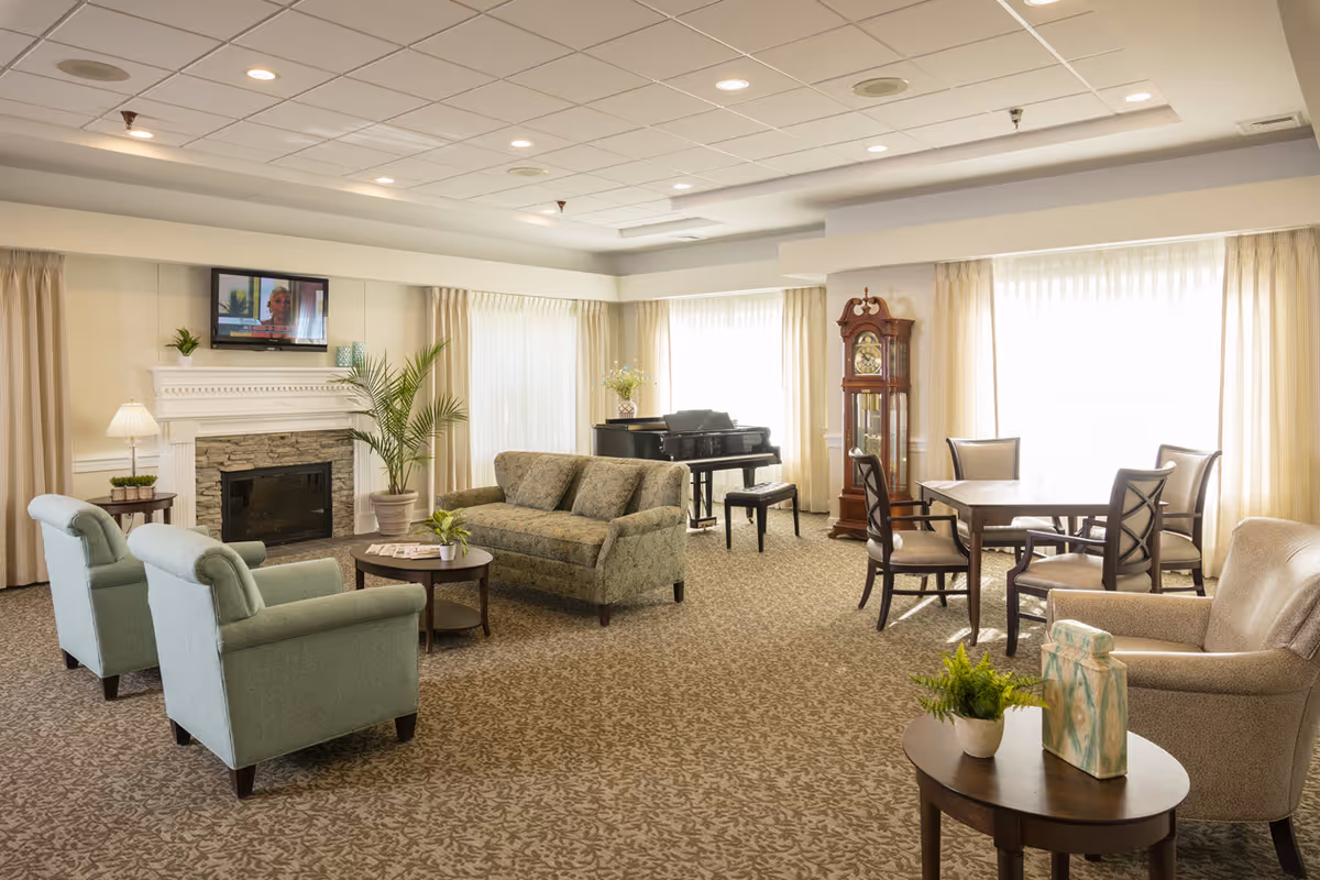 Bright communal lounge with sofas and armchairs arranged around a fireplace, a piano, a grandfather clock and a dining table by large windows.