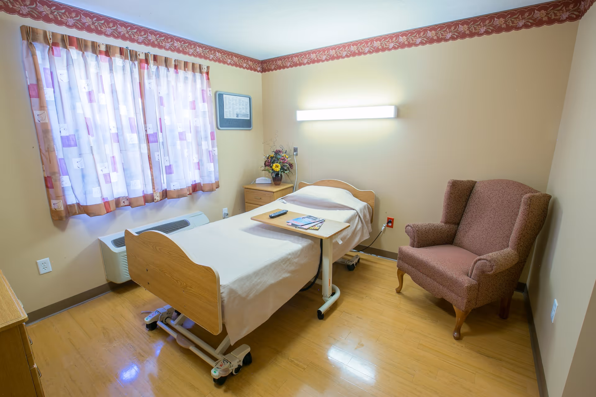 A small nursing facility bedroom with a single hospital-style bed covered with a white sheet, a wooden bedside table with a flower arrangement and a telephone, a pink upholstered armchair, and a window with patterned curtains letting in natural light. The room has light-colored walls with a floral border near the ceiling and a wooden floor.