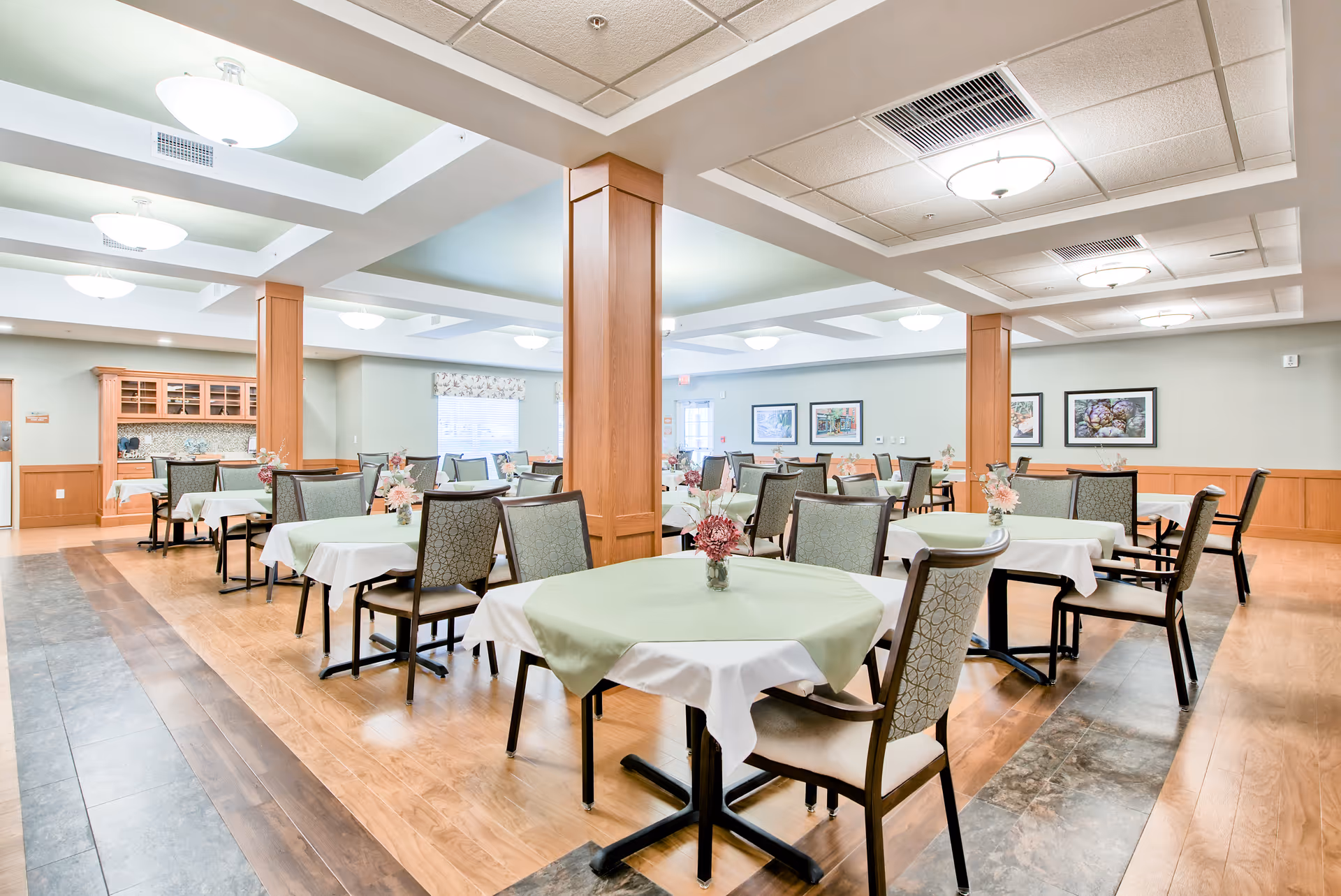A spacious dining room with multiple tables covered with white and light green tablecloths, each adorned with a small floral centerpiece. The room features wooden flooring with a decorative strip, wooden pillars, soft green walls, ceiling lights, and framed artwork on the walls.