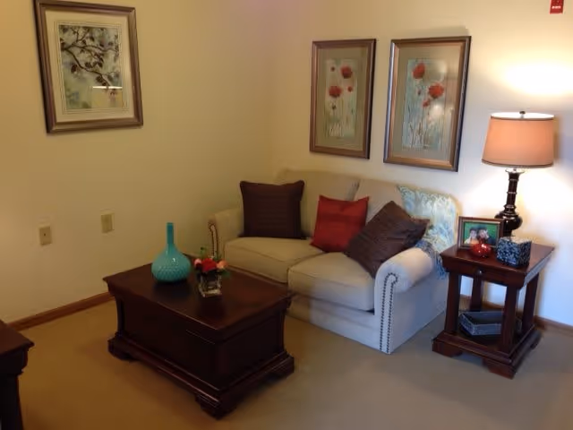 A cozy living room area with a beige loveseat adorned with three decorative pillows in red and brown. Above the loveseat are two framed floral paintings. To the right of the loveseat is a wooden side table with a lamp, a framed photo, and decorative items. In front of the loveseat is a wooden coffee table with a turquoise vase and a small flower arrangement. The walls are light-colored and the floor is carpeted.