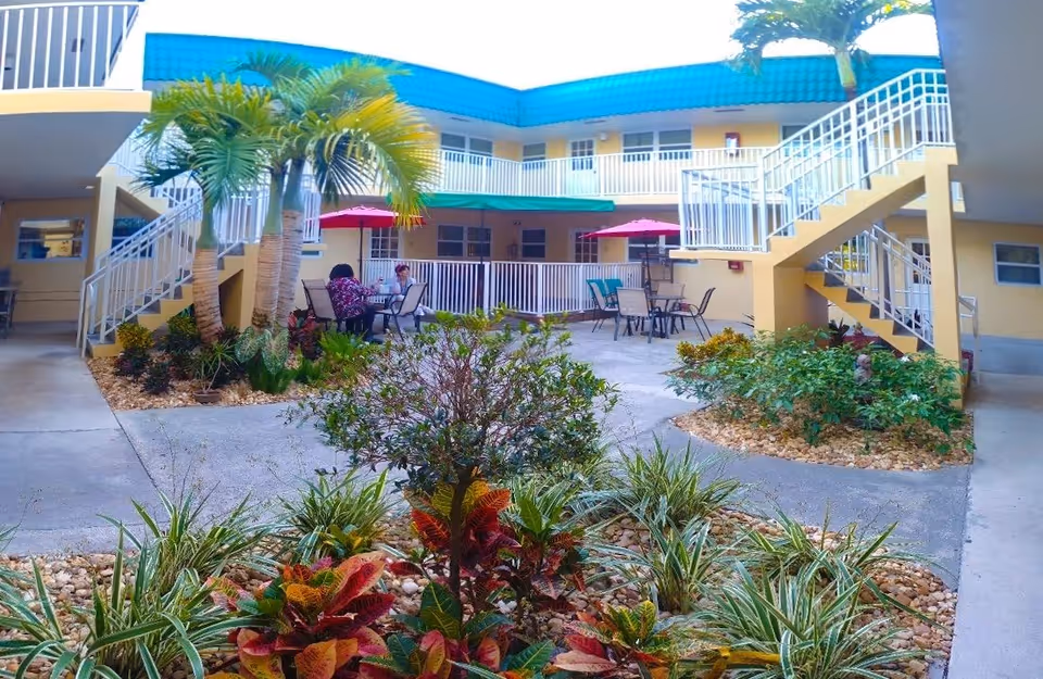 Outdoor courtyard area of a senior living facility with tropical plants and shrubs in landscaped beds. There are two palm trees near the center, several tables with chairs and red umbrellas where two people are seated. The building surrounding the courtyard is two stories with yellow walls, white railings, and a blue roof. Stairs lead to the upper floor on both sides of the courtyard.