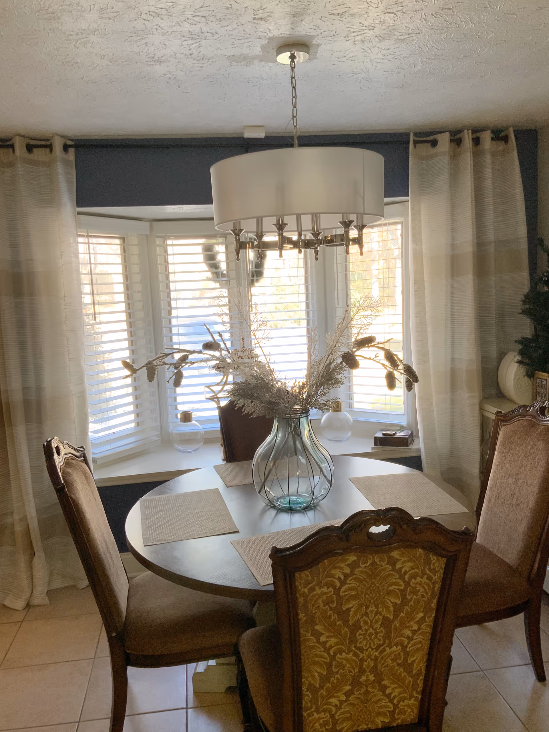 A dining area with a round wooden table set with four placemats and a decorative glass vase with dried floral arrangement in the center. The table is surrounded by four upholstered chairs with ornate wooden backs. Behind the table is a bay window with white blinds and beige curtains. A modern chandelier with a white drum shade hangs above the table.