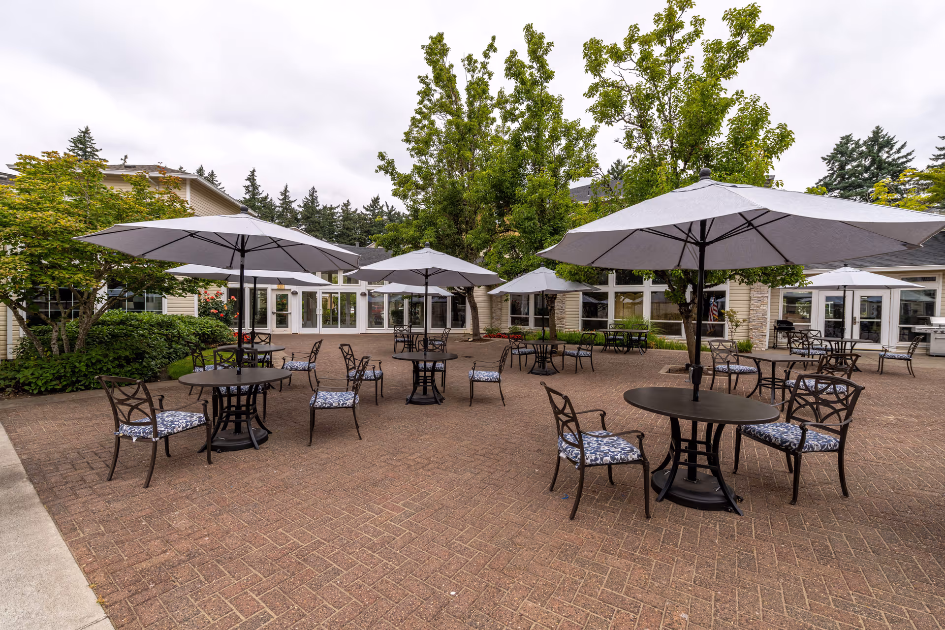Outdoor patio area with multiple round tables and chairs, each table shaded by large white umbrellas. The patio is paved with brick and surrounded by greenery and trees, with a building visible in the background under a cloudy sky.