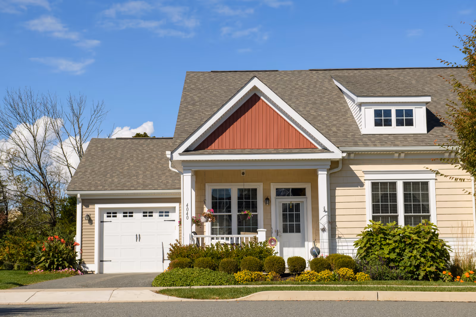 Front exterior of a light-colored house with an attached garage, covered porch, dormer window, and landscaped shrubs under a blue sky.
